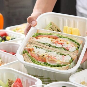 A person holds a plastic container with sandwiches filled with lettuce, tomato, and cabbage. The background shows other food containers, including rice dishes, watermelon slices, and salad. A yellow bell pepper is visible in the corner.