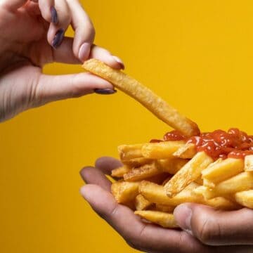 A hand holds a french fry dipped in ketchup from a pile of fries cupped in another hand against a solid yellow background.
