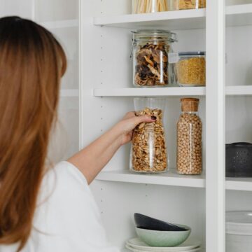 A person with long brown hair is organizing a white pantry shelf stocked with essential vegan pantry ingredients. The shelf boasts jars filled with pasta, nuts, and grains, alongside mugs and bowls. The person is holding a jar of dried fruit.
