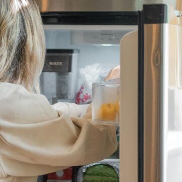 A woman with long blonde hair is reaching into an open refrigerator. The fridge contains various items, including a visible watermelon. The kitchen has natural light coming from a nearby window with curtains.