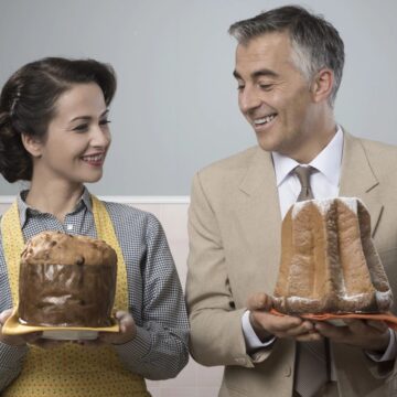 A woman and a man stand side by side, each holding a delightful pastry. The woman displays a round, dark brown cake, while the man reveals a tall confection dusted with powdered sugar. Both are smiling, seemingly admiring each other's selections like an impeccable auto draft of delectable choices.