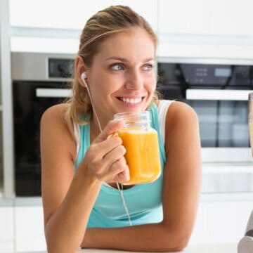 A person sitting in a kitchen holds a glass mug containing an orange smoothie. A blender with more smoothie is on the counter. The person is wearing earbuds and smiling, and the background features shelves and modern kitchen appliances.