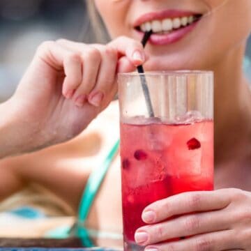 A person leans on the edge of a pool, holding a glass with a red drink filled with ice and berries. They are sipping from a black straw and wearing a turquoise swimsuit. The water in the pool is visible in the background.