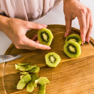 A person is slicing kiwis on a round wooden cutting board. Several peeled kiwi slices are positioned next to a pile of green peels. A knife rests on the board to the left of the person's hands.