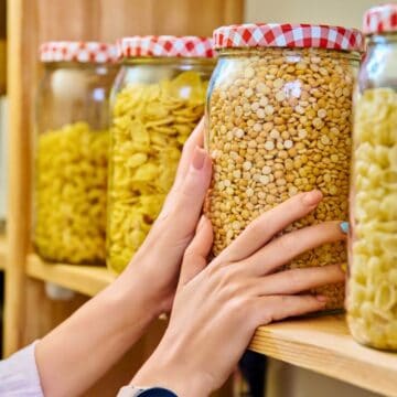 Hands are arranging glass jars filled with pasta and legumes on a wooden pantry shelf. The jars have red and white checkered lids. Other shelves with kitchen items are visible in the background.