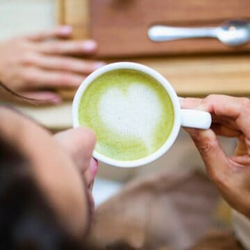 A person is holding a cup of matcha latte with latte art resembling a heart. The scene is from above, showing part of their face and hands. A wooden tray with a spoon is visible in the background on a table.