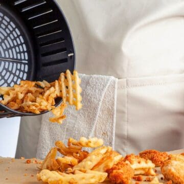 A person in a white apron is pouring freshly cooked waffle fries onto a piece of parchment paper. Beside the fries are several pieces of breaded chicken. The food is being transferred from the basket of an air fryer.