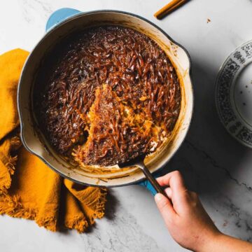 A round baking dish with a brown baked dessert, partially served, sits on a marble countertop. A hand holds the dish with an orange cloth napkin nearby. A fork rests on a patterned plate with crumbs. Cinnamon sticks are scattered around, hinting at the delicious recipe for Jerusalem Kugel.
