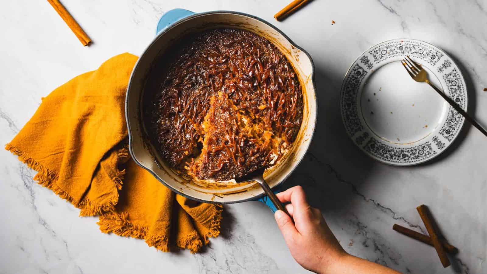 A round baking dish with a brown baked dessert, partially served, sits on a marble countertop. A hand holds the dish with an orange cloth napkin nearby. A fork rests on a patterned plate with crumbs. Cinnamon sticks are scattered around, hinting at the delicious recipe for Jerusalem Kugel.