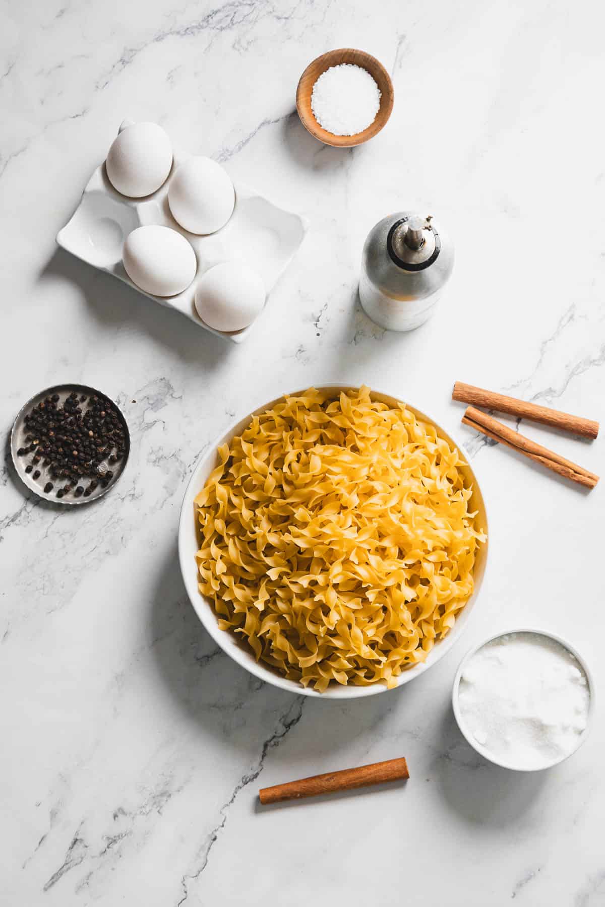 A white bowl filled with uncooked egg noodles is placed on a marble surface next to a plate with whole black peppercorns, a tray of four eggs, a small bowl of salt, a pepper grinder, cinnamon sticks, and a bowl of flour.