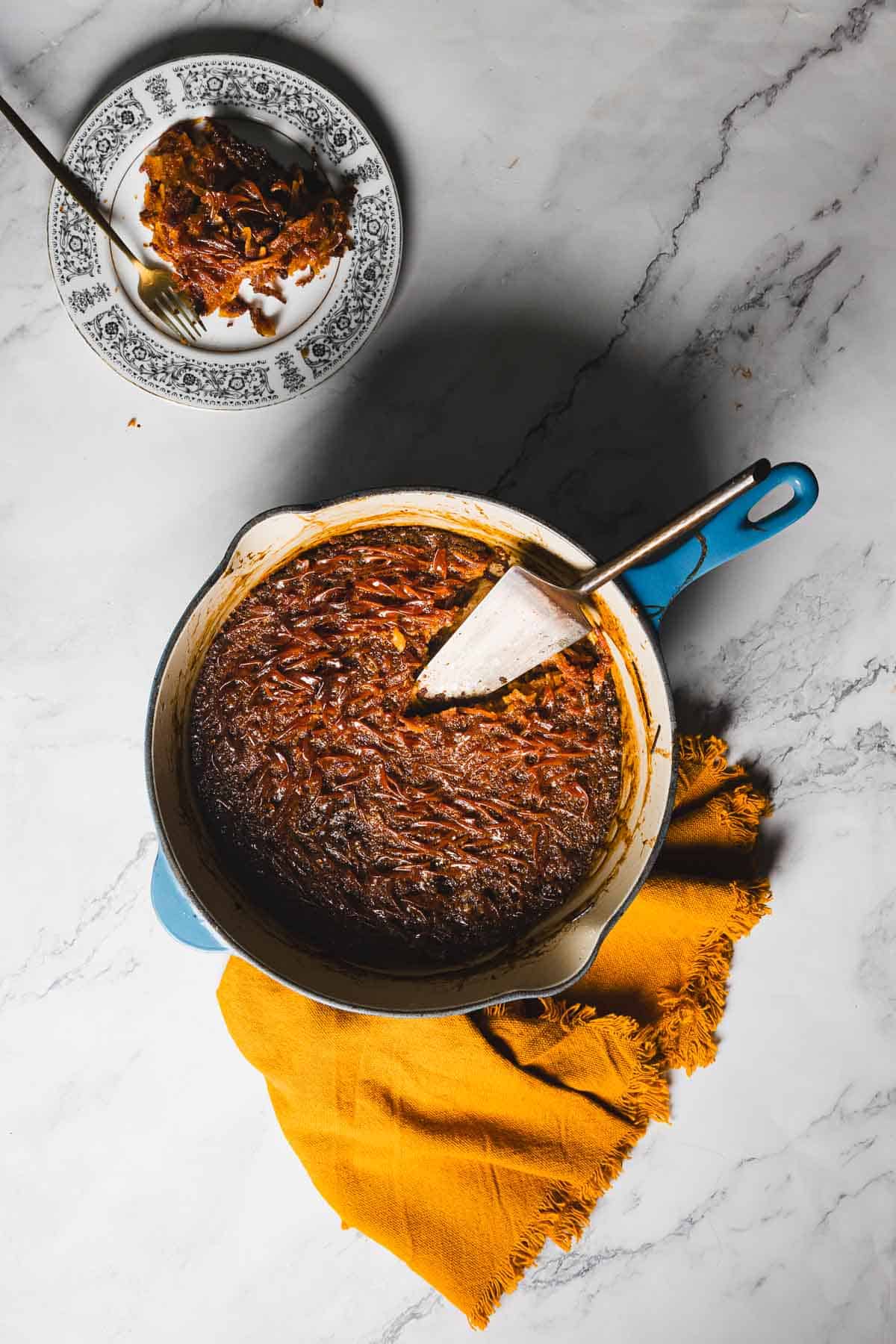 A blue pot filled with cooked stew is placed on a marble countertop with a yellow cloth underneath. Beside the pot is a small plate with a portion of the stew and a fork. The plate has a black and white decorative pattern.