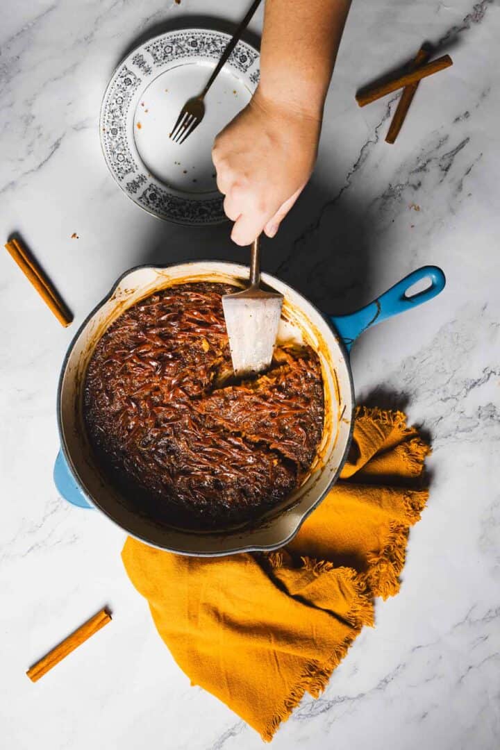 A hand using a spatula to serve food from a blue-handled skillet filled with a baked dish. The skillet rests on a marble surface near a yellow cloth, a patterned plate, a fork, and a few scattered cinnamon sticks.