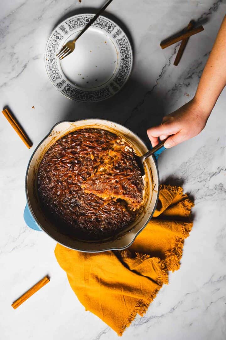 A person's hand is scooping from a round, blue pot of cooked food, likely a stew or casserole, placed on a marble countertop with a yellow cloth underneath. A patterned, empty plate with a fork and scattered cinnamon sticks are nearby.