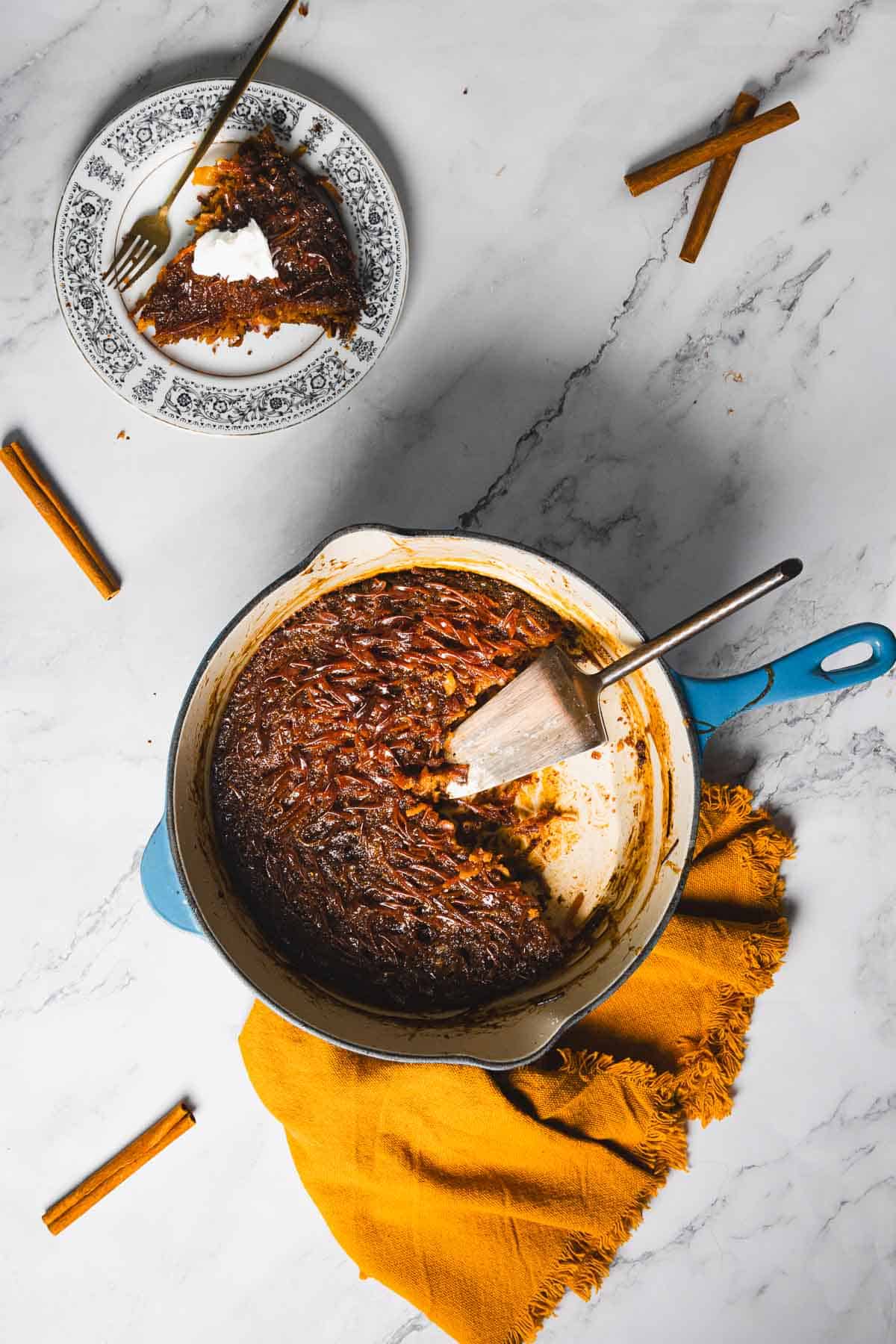 A cast iron skillet containing partially sliced dessert sits on a white marble surface with a yellow cloth underneath. A slice of the dessert is served on a patterned plate nearby, accompanied by a fork. Cinnamon sticks are scattered around the setup.