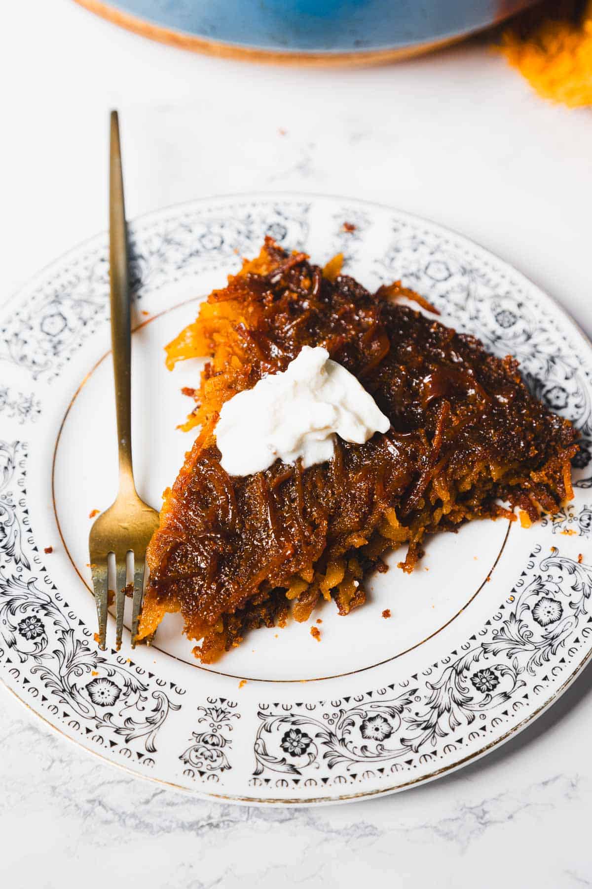 A slice of brown pie with a dollop of white cream on top is placed on a decorative plate, accompanied by a gold fork. The pie has a caramelized texture and the cream adds a contrasting color. The plate rests on a white surface.