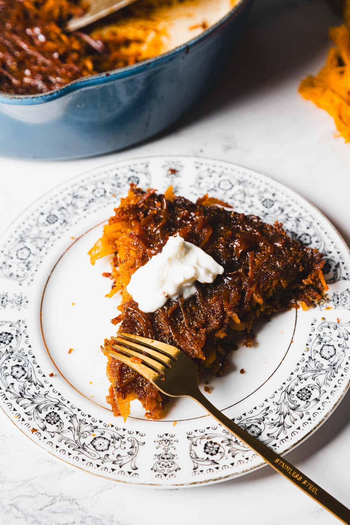 A slice of baked dessert topped with a dollop of cream on a decorative white plate with black floral designs. A gold fork rests on the edge of the plate. The background shows a baking dish with more dessert.