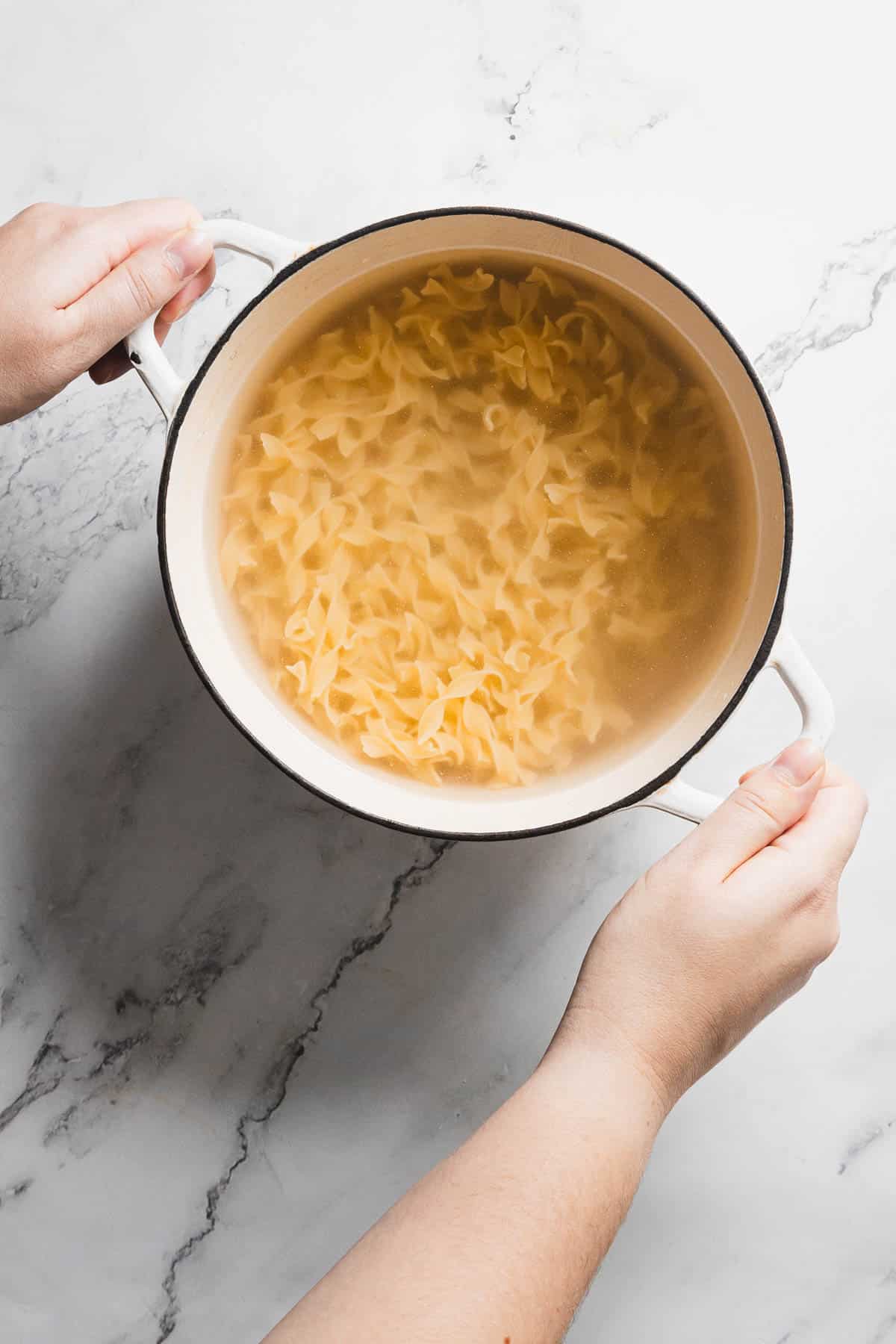 A person is holding a white pot filled with cooked noodles submerged in water. The pot is positioned against a marble countertop. The person's hands are gripping the pot handles on each side.