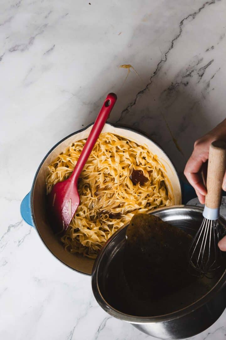 A person stands near a pot of cooked noodles with a red spatula resting on the noodles. The person holds a metal bowl and a whisk, appearing to pour liquid from the bowl into the pot of noodles on a marble surface.