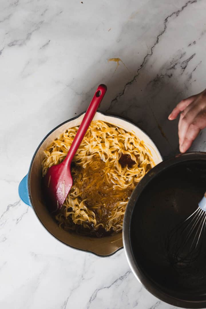 A person holds a pot above a blue cast-iron pot filled with fettuccine noodles and a sauce, stirring with a red spatula. The scene is set on a white marble countertop.