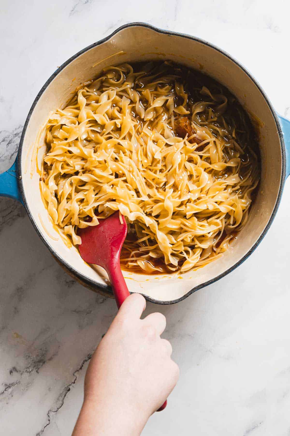 A hand stirring cooked noodles in a blue pot with a red spatula. The noodles appear to be coated in a sauce, and the scene is set on a white marble surface.