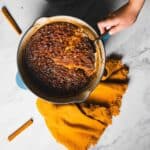 A hand is holding a spoon above a round baking dish filled with a baked dessert that has a caramelized, golden top. A yellow fringed cloth is placed beside the dish on a marble surface, with two cinnamon sticks lying nearby.