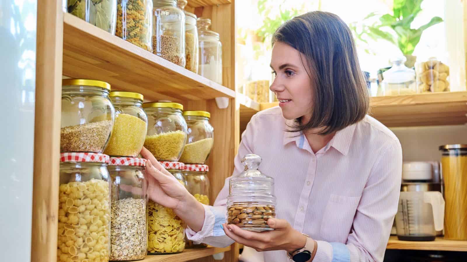 A woman in a pink shirt is organizing glass jars filled with various groceries, such as pasta and grains, on wooden shelves. She holds a jar of cookies in her hand. The background includes potted plants and kitchen appliances.