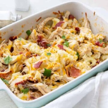 A casserole dish filled with a baked mixture of shredded chicken, melted cheese, bacon pieces, and garnished with parsley. A pepper shaker is in the background, and a white cloth lays beside the dish.
