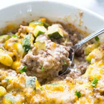 A close-up of a bowl containing a creamy zucchini and ground beef casserole. The dish includes melted cheese and is garnished with herbs. A spoon is placed in the casserole.