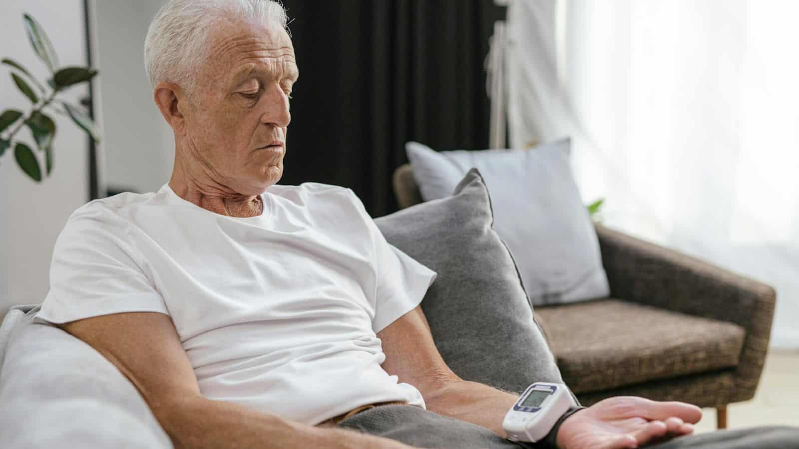 An elderly man with white hair is sitting on a couch, wearing a white t-shirt, and monitoring his blood pressure with a digital blood pressure device strapped to his wrist. He appears focused on the results. A plant, an armchair, and curtains are in the background.