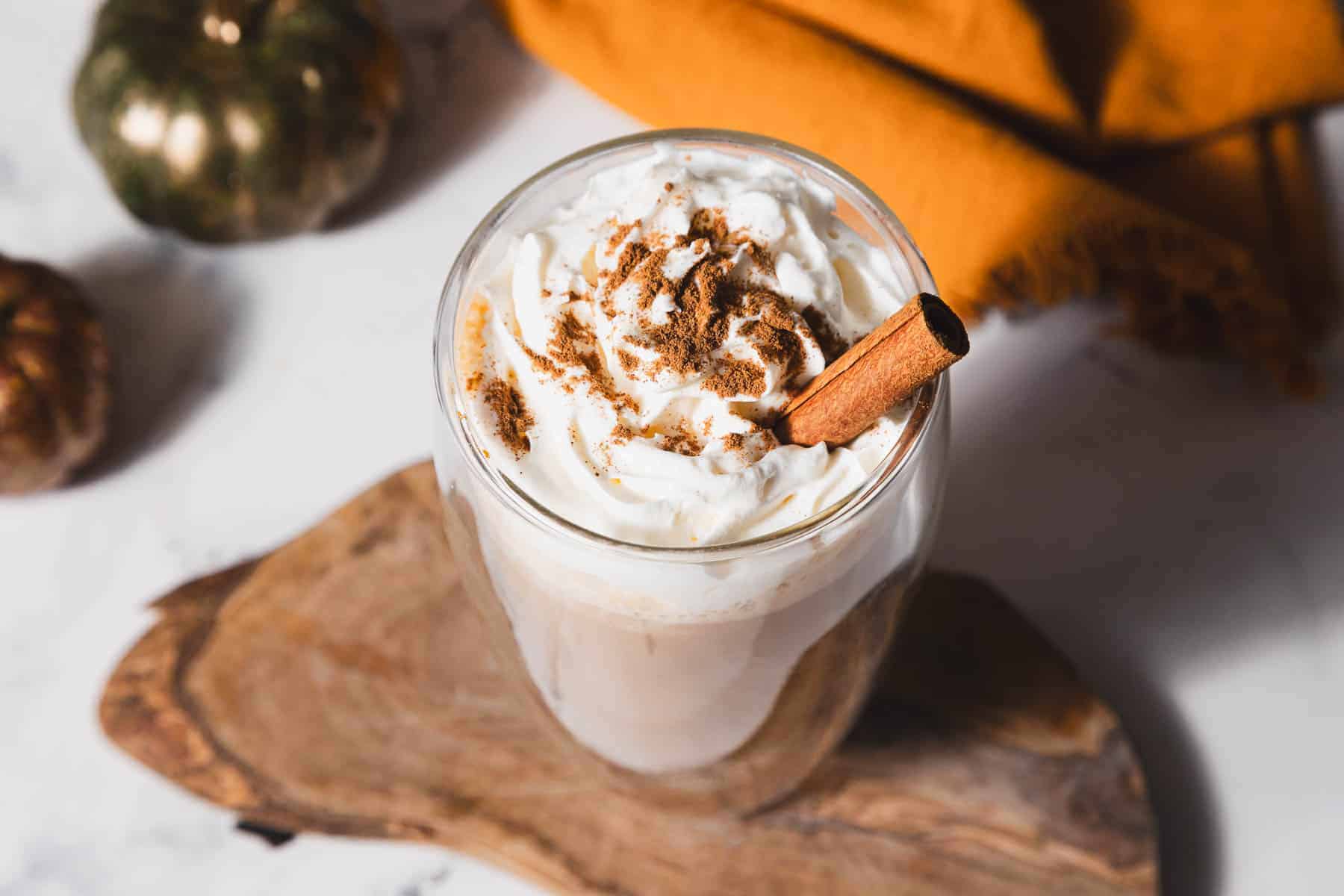 A glass mug filled with a pumpkin spice latte topped with whipped cream, sprinkled with cinnamon, and garnished with a cinnamon stick. The mug is on a wooden board, with a small pumpkin and an orange cloth in the background.