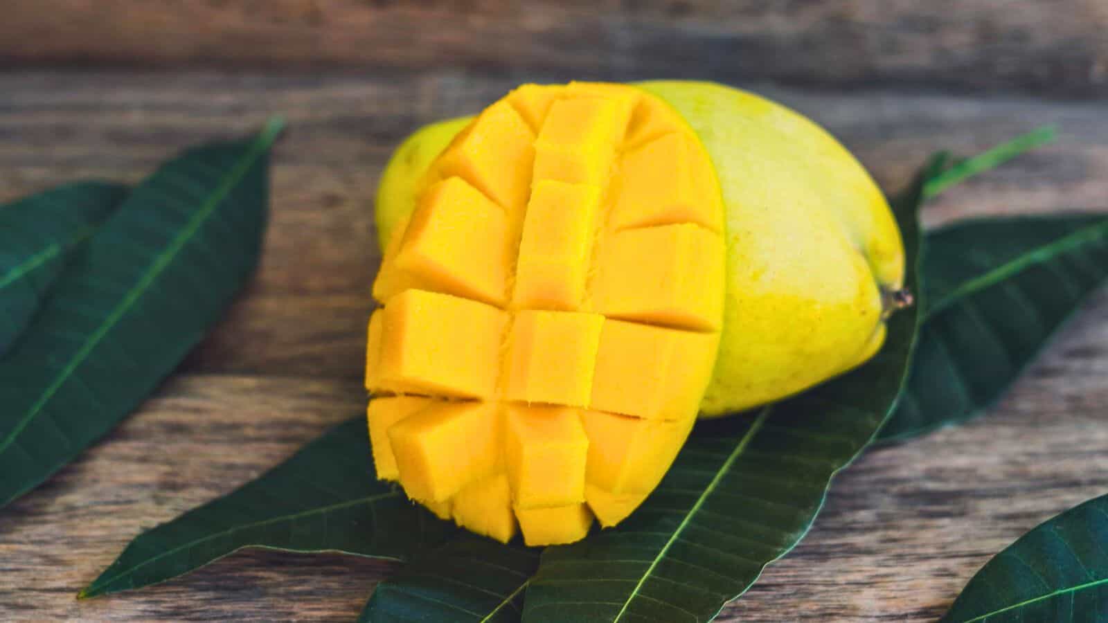 A ripe mango is sliced into cubes and placed on a wooden surface, with a whole mango and green leaves in the background.