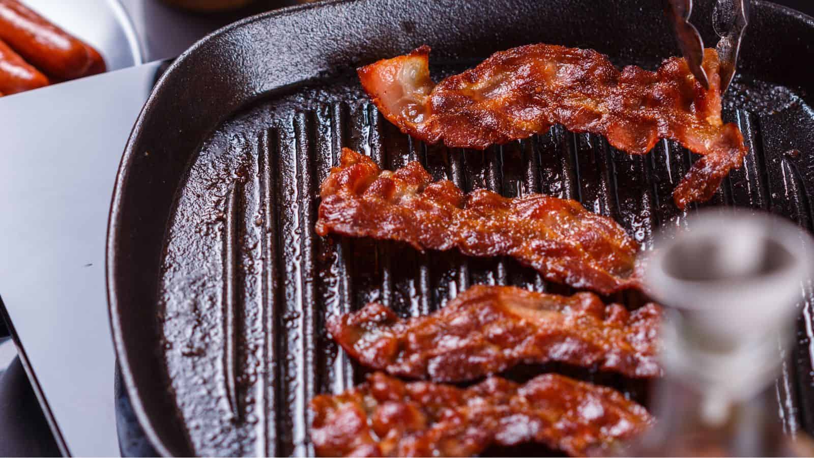 Close-up of crispy bacon strips being cooked on a black ridged grill pan. A pair of metal tongs is used to handle the bacon. In the background, there are sausages on a plate.