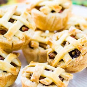 A plate holds several mini apple pies with golden, flaky lattice crusts. They are arranged closely together, showcasing the baked filling through the lattices. The background is blurred with colorful fabric.