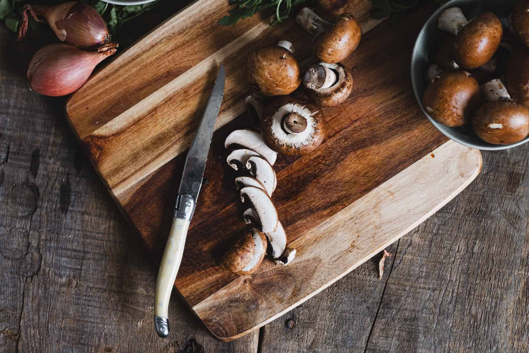 A wooden cutting board with a knife and sliced brown mushrooms. Whole mushrooms and shallots are also on the board. A bowl with more whole mushrooms is nearby on a rustic wooden surface.