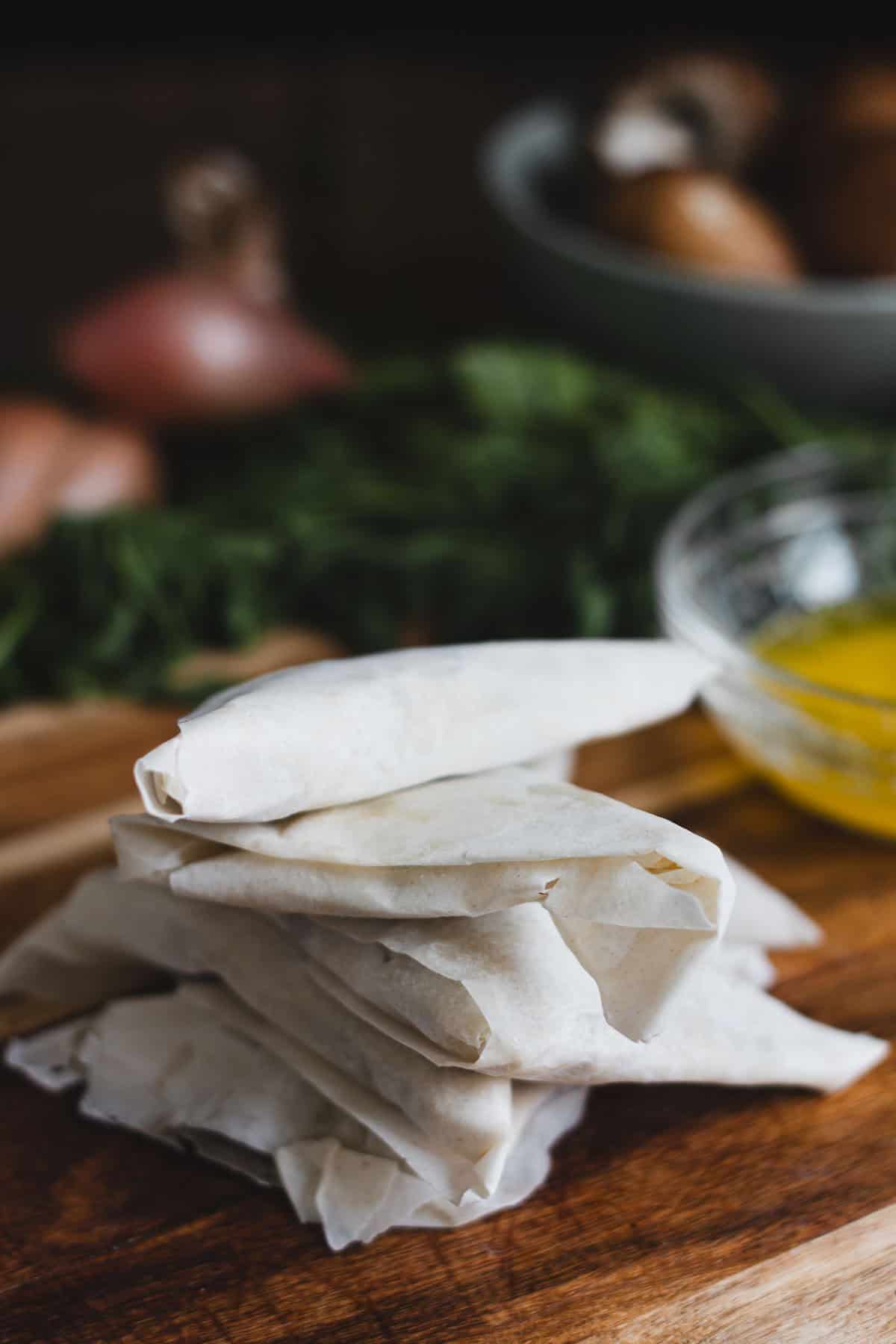 A stack of rolled pastry sheets sits on a wooden cutting board, with a glass bowl of melted butter nearby. In the background, there are green herbs and vegetables slightly out of focus.