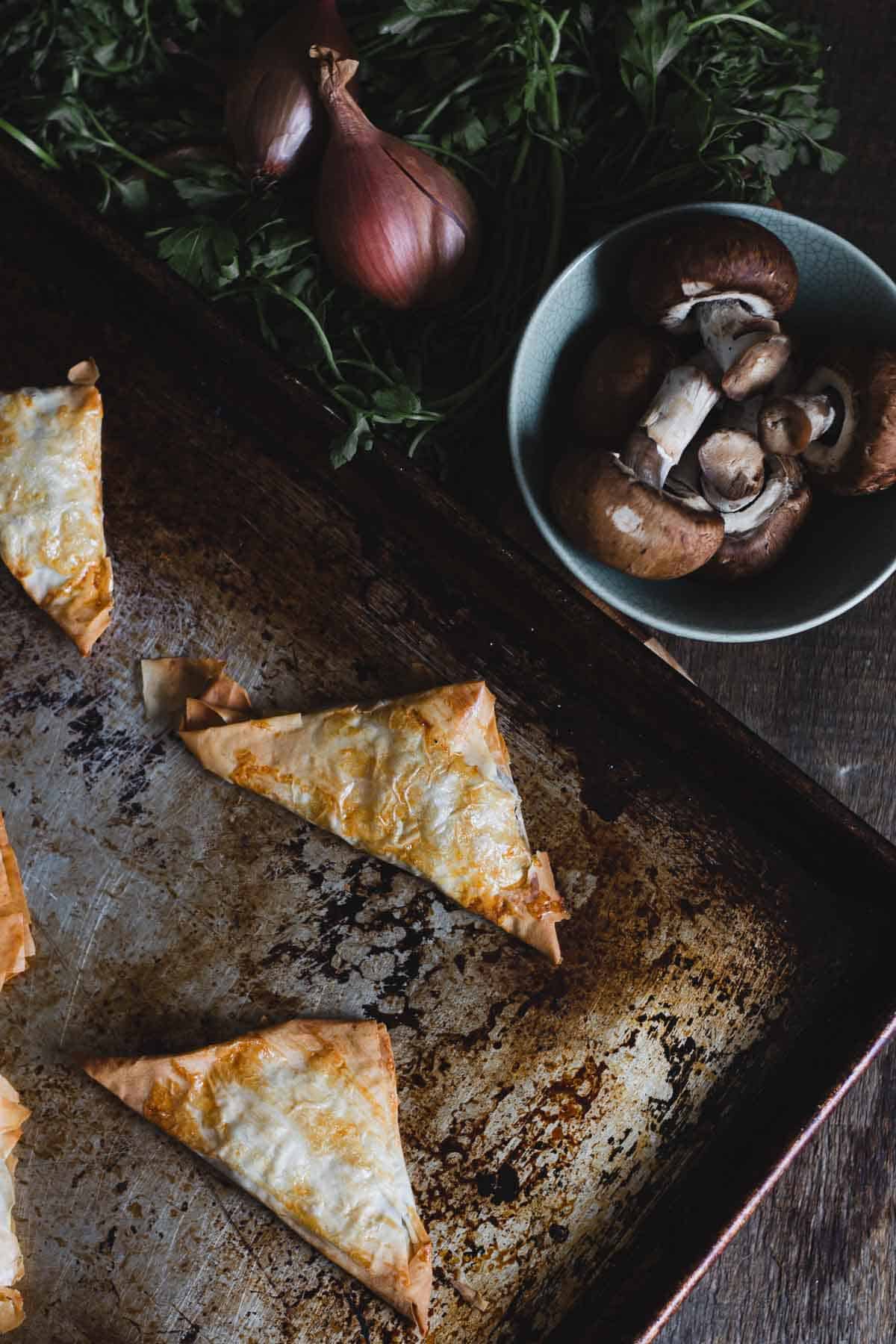 A baking tray with three golden-brown triangular pastries is shown. In the background, a bowl of mushrooms and three whole shallots rest on a wooden surface. Fresh herbs are partially visible.