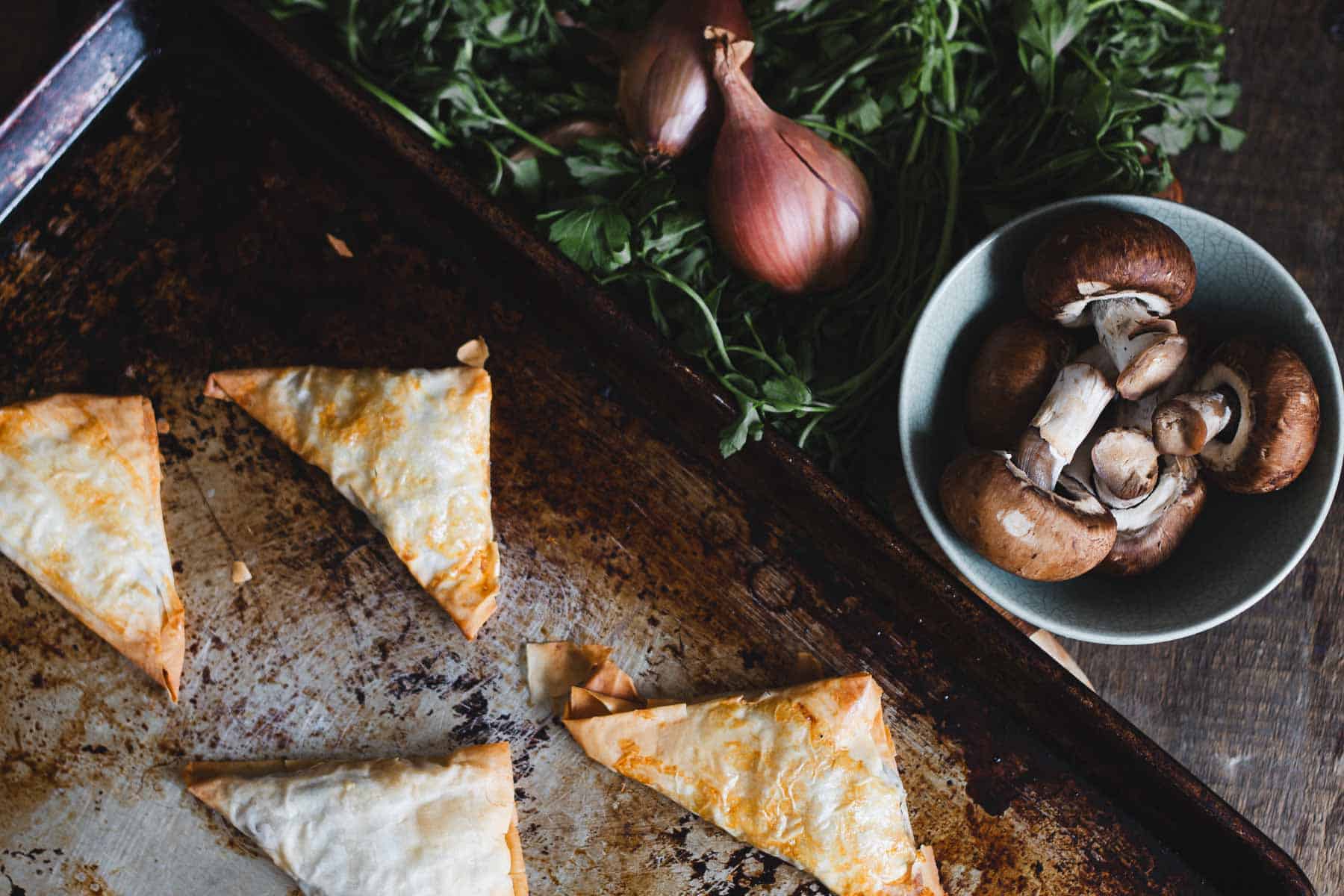 Golden-brown pastry triangles are on a baking tray. Beside the tray, there are shallots and leafy greens on a dark surface, and a bowl filled with mushrooms.