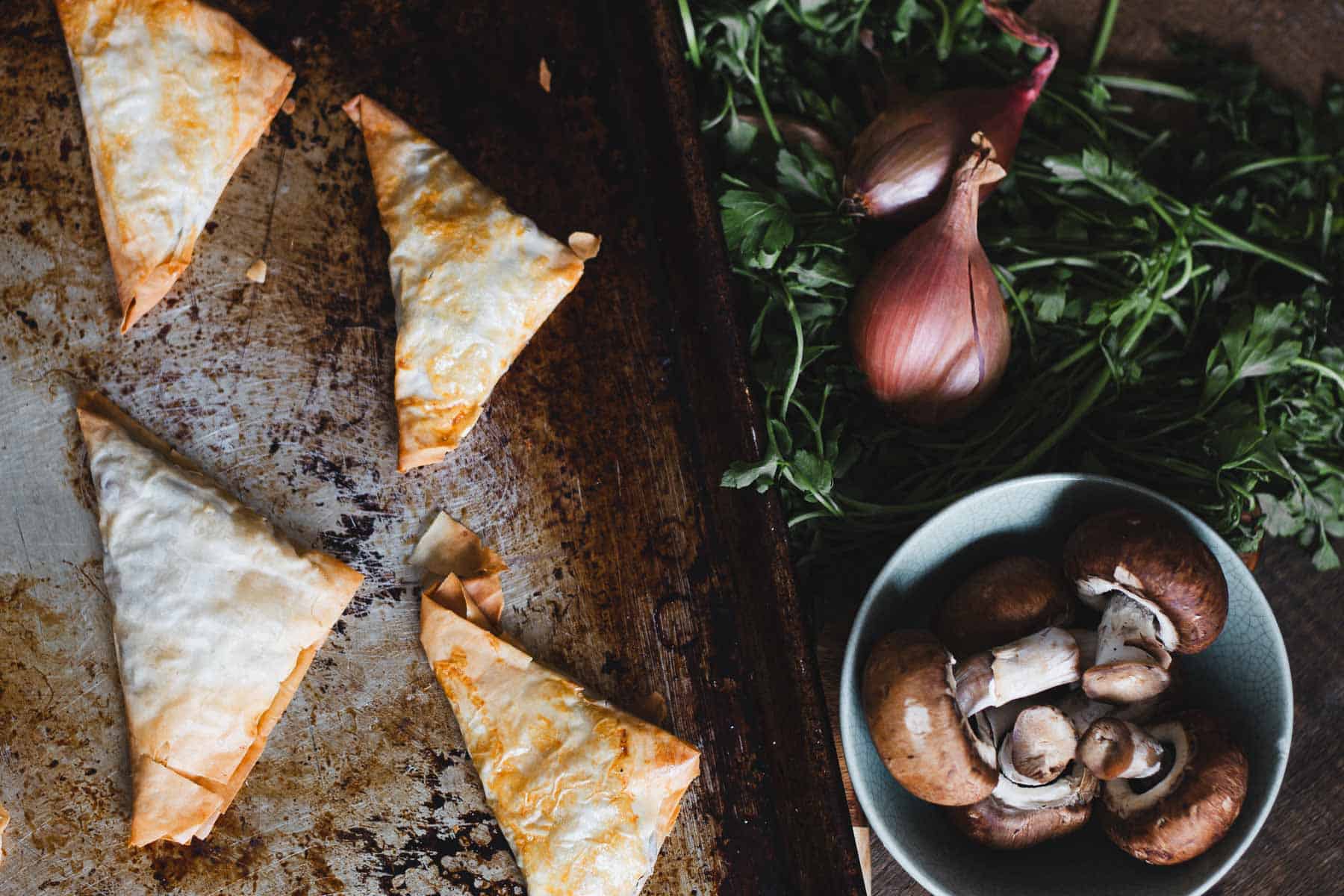 Four triangular pastries are on a baking tray. To the right, a cluster of fresh herbs lies next to two whole shallots. A bowl filled with whole mushrooms is in the bottom right corner of the image.