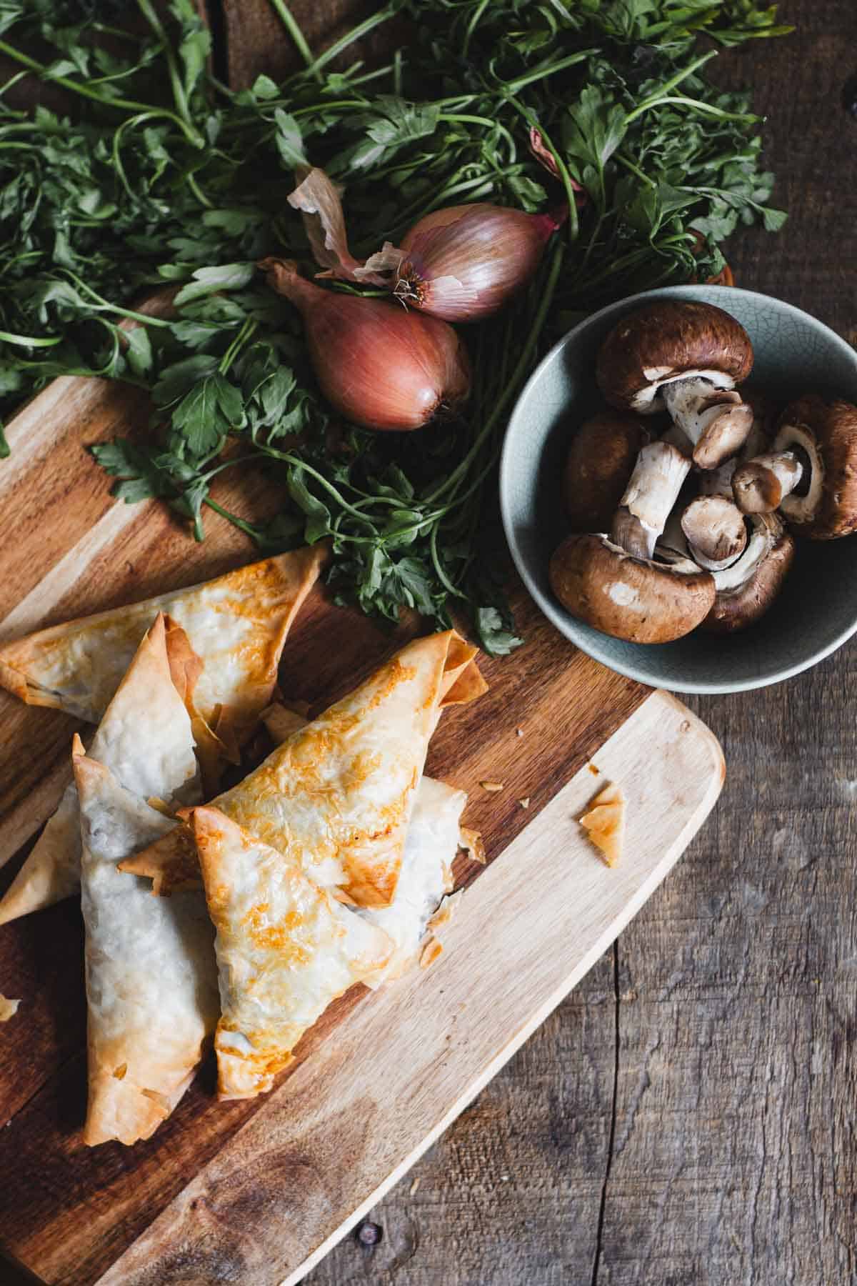 A wooden board with golden-brown baked pastries, surrounded by fresh herbs and shallots. A small bowl filled with whole and sliced mushrooms is nearby. The scene is set on a rustic wooden surface.