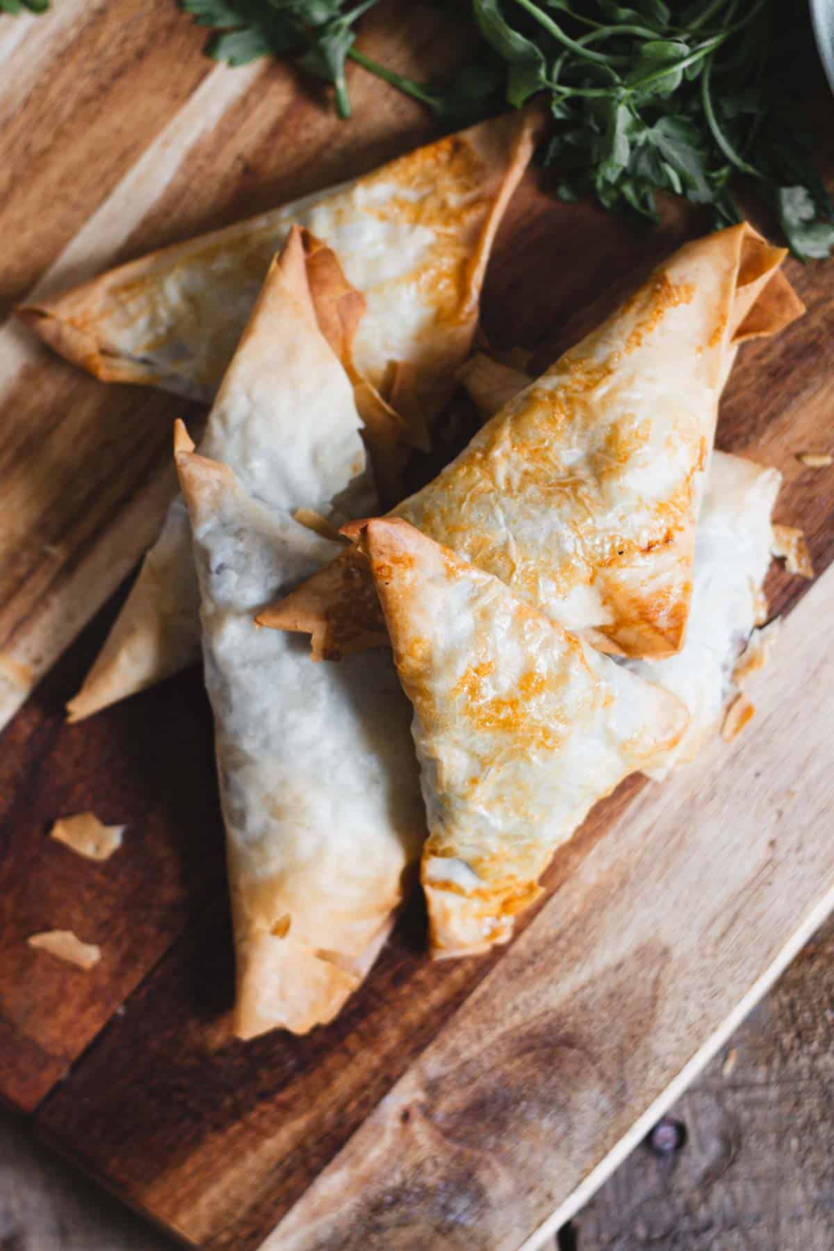 Five golden-brown triangular pastries are arranged on a wooden cutting board, with some herbs partially visible in the background.