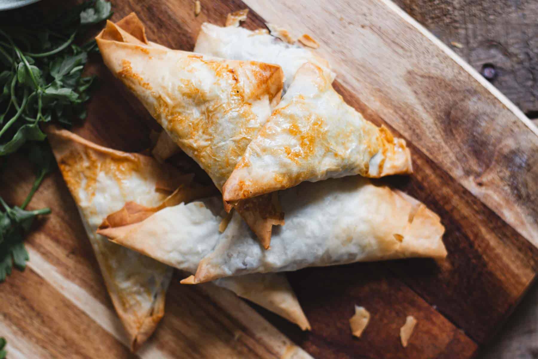 A stack of golden-brown, triangular pastries on a wooden cutting board. The pastries have a flaky texture and are surrounded by fresh parsley leaves.