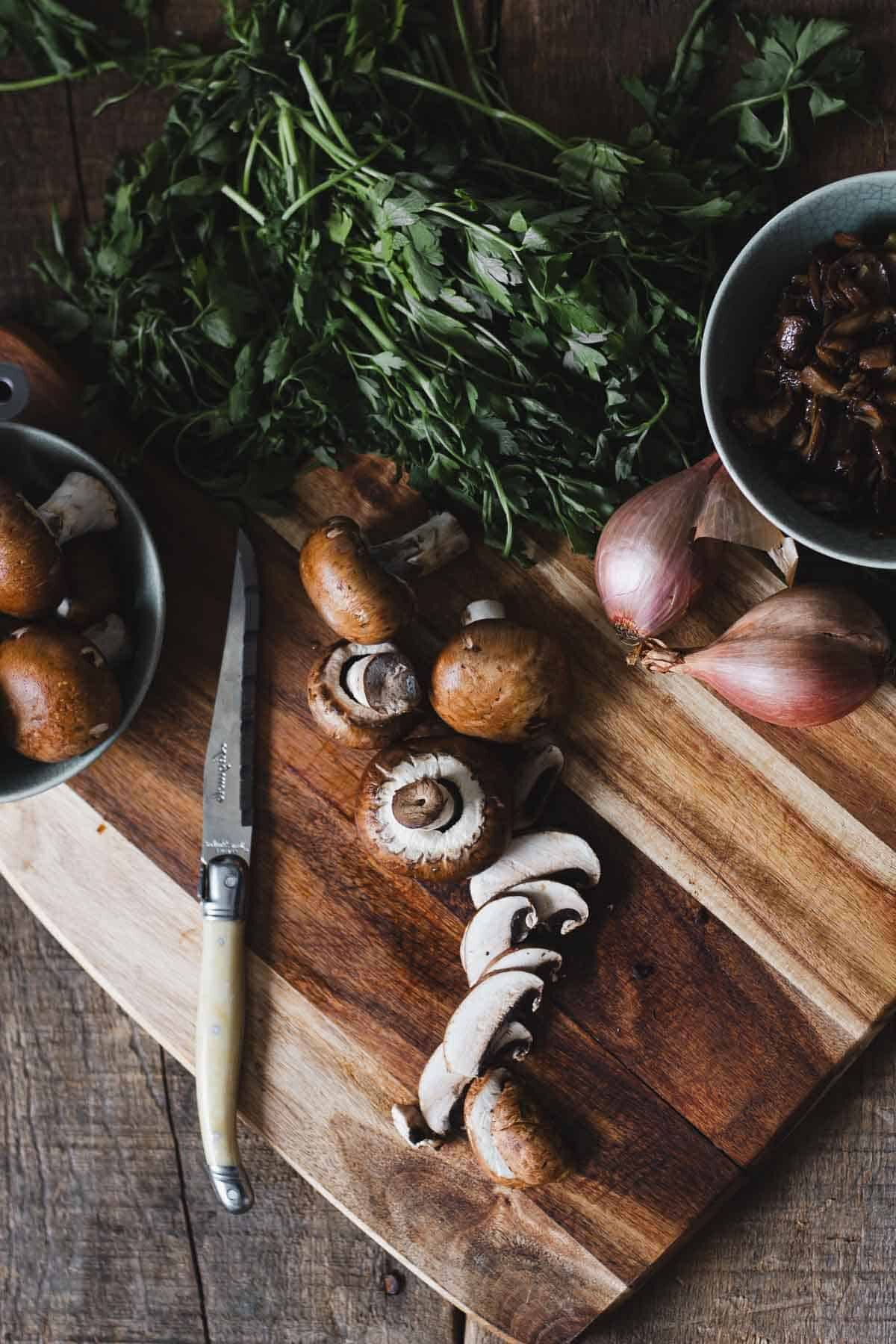 A wooden cutting board with sliced mushrooms, whole mushrooms, fresh parsley, and two shallots. A knife is placed nearby. Two bowls contain more mushrooms and chopped parsley. The board is on a rustic wooden surface.