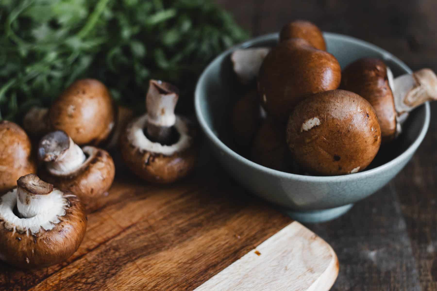 A group of brown mushrooms are on a wooden cutting board and in a gray bowl. Fresh green herbs are partially visible in the background. The setting is on a dark wooden surface.