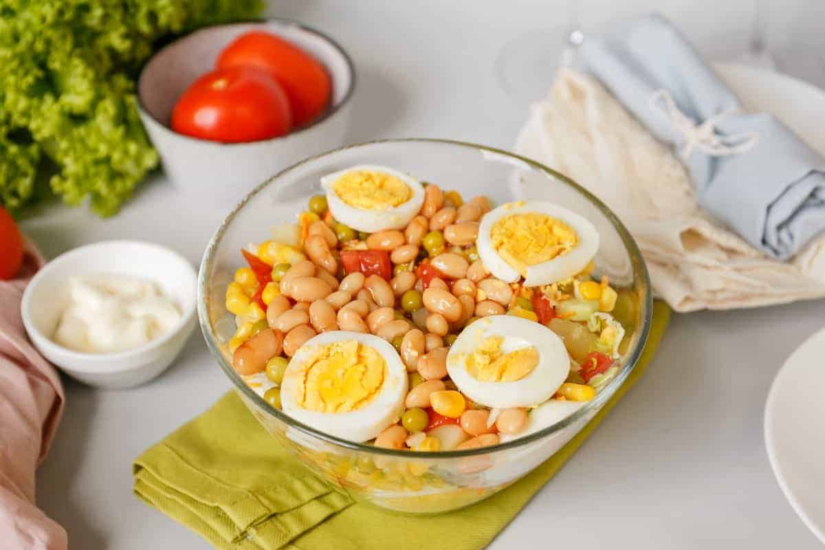 A glass bowl is filled with a salad of white beans, corn, and sliced boiled eggs. The setting features fresh tomatoes, lettuce, a small bowl of mayonnaise, and a stack of folded napkins&mdash;a delightful scene for discovering favorite recipes.