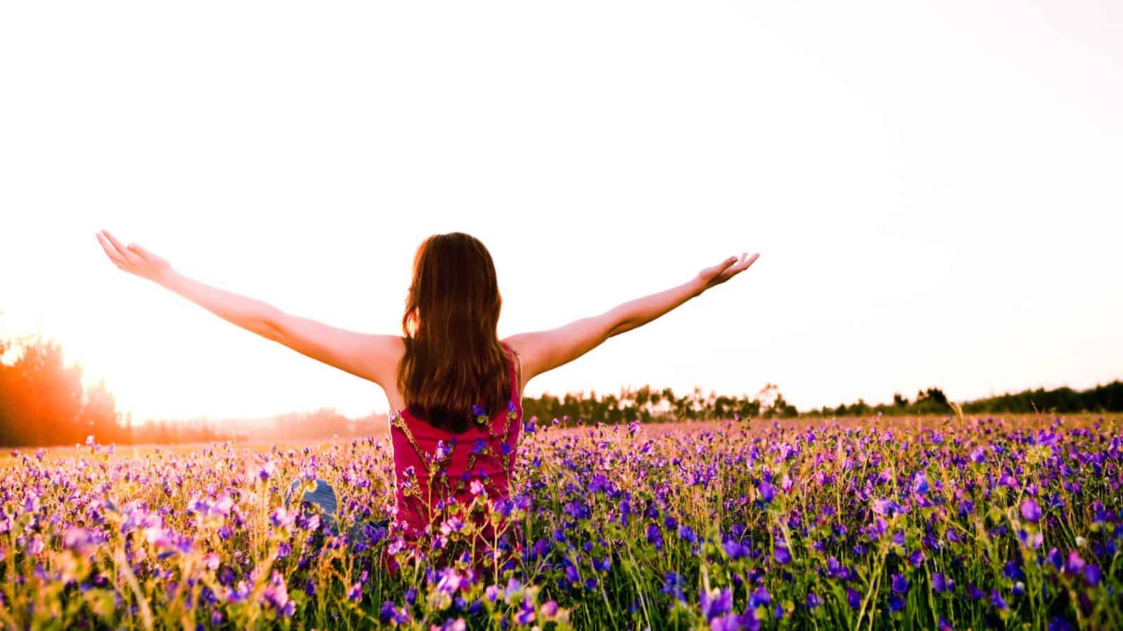 A person with long hair, wearing a sleeveless outfit, sits in a field of purple and yellow flowers with their back to the camera, arms outstretched. The sun is setting or rising on the horizon, casting a warm glow over the scene.