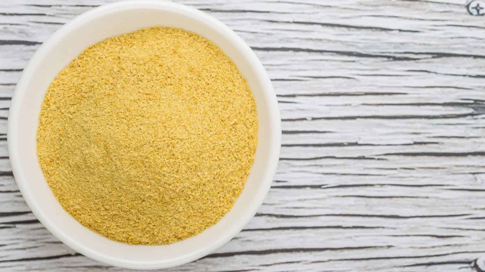 A white bowl filled with yellow nutritional yeast flakes is placed on a wooden surface with a light and dark wood grain pattern.