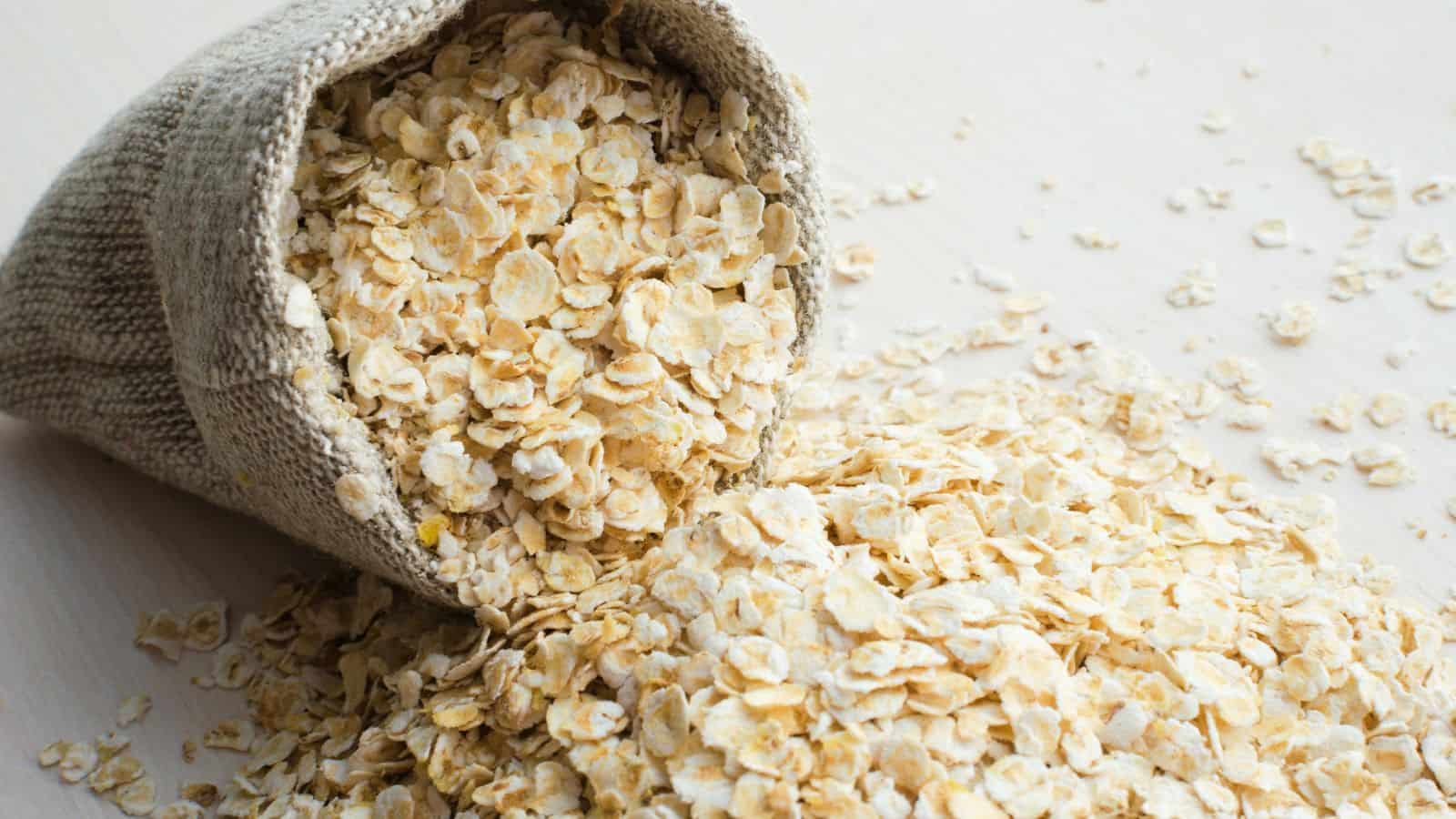 A burlap sack is tipped over on a light wooden surface, spilling a pile of rolled oats onto the table. The oats are scattered around the sack, creating a textured surface.