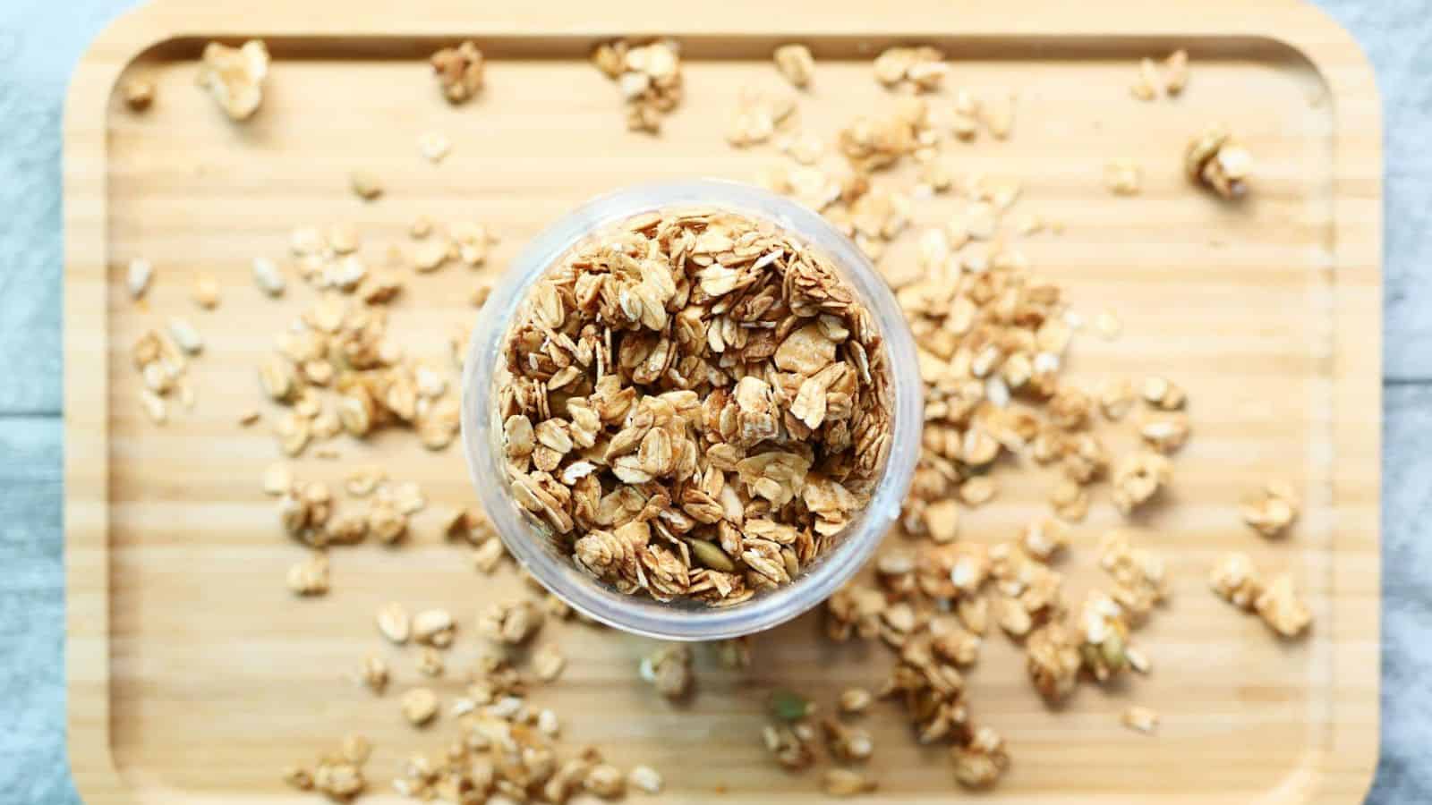 A glass jar filled with granola sits on a wooden cutting board. Some granola pieces are scattered around the jar. The cutting board rests on a light gray surface.