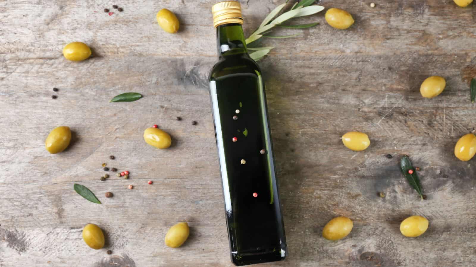 A dark glass bottle with a gold cap lies on a wooden surface surrounded by green olives and scattered peppercorns. An olive branch with leaves is placed near the bottle, adding a touch of greenery to the arrangement.