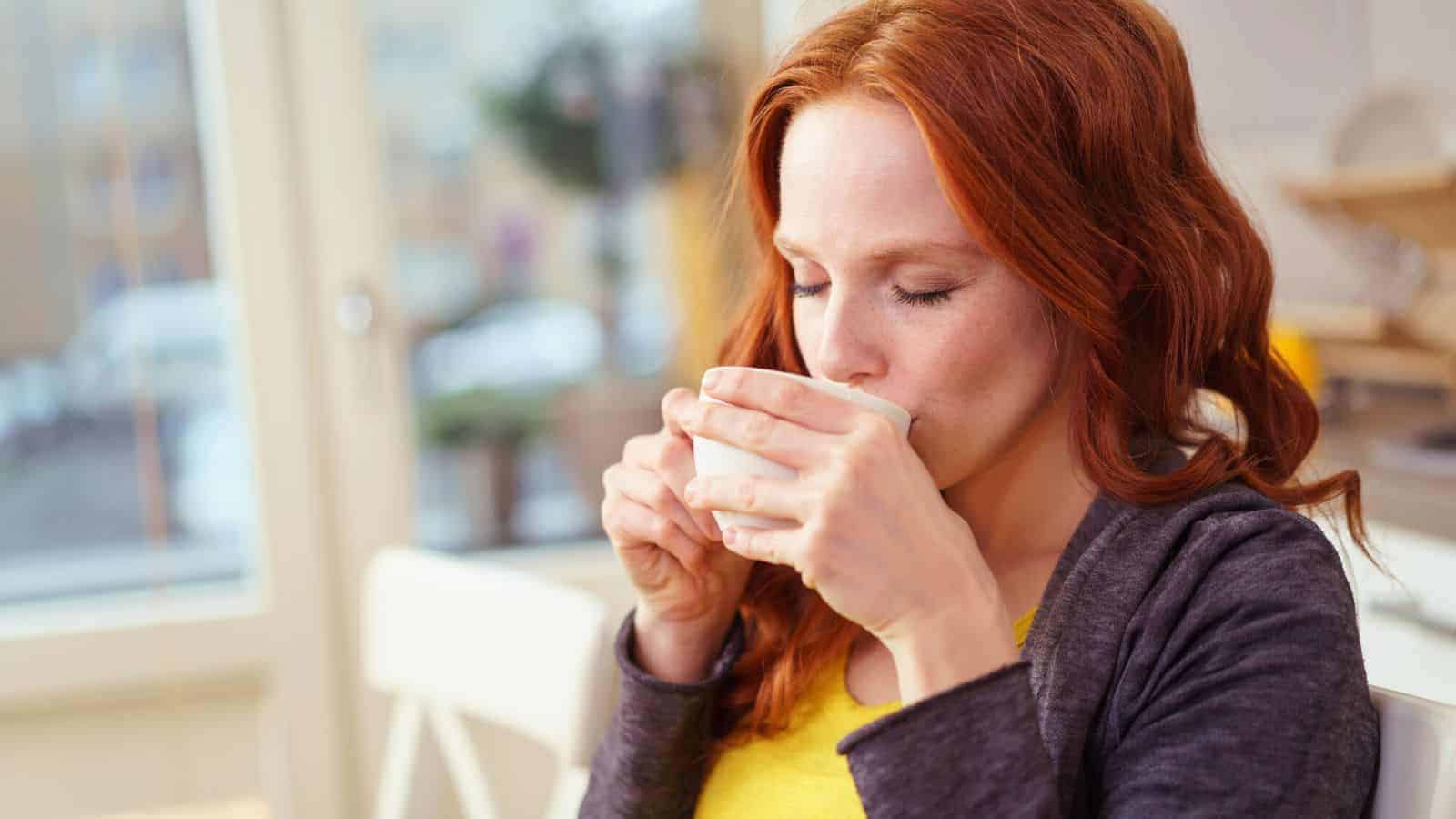 A woman with long red hair sips from a white cup in a bright room. She is wearing a gray sweater over a yellow shirt and appears to be enjoying a warm beverage. The background shows a window and blurred interior details.